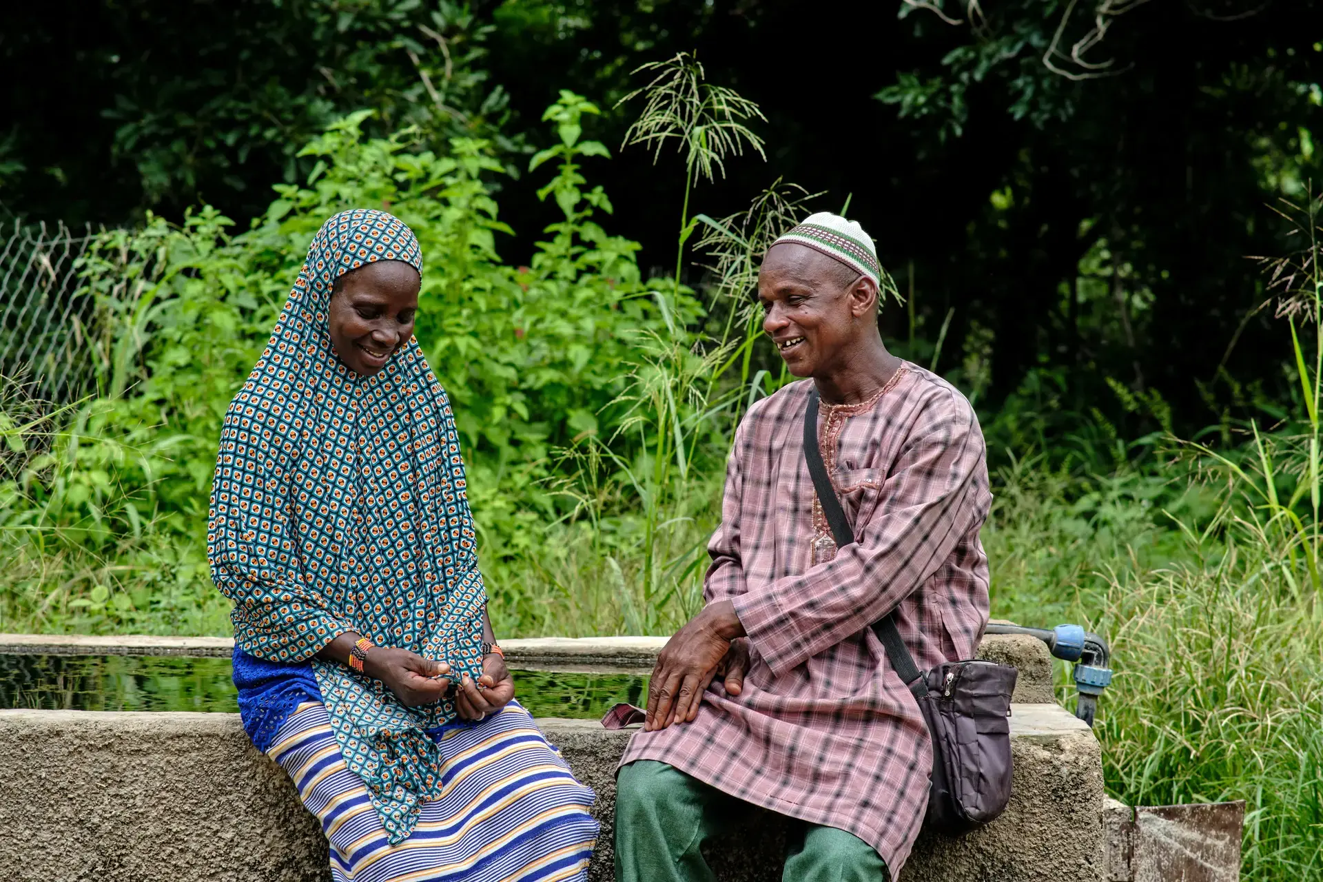 A man and woman wearing traditional clothing, sitting side by side on a water container outside in natural setting, smiling.